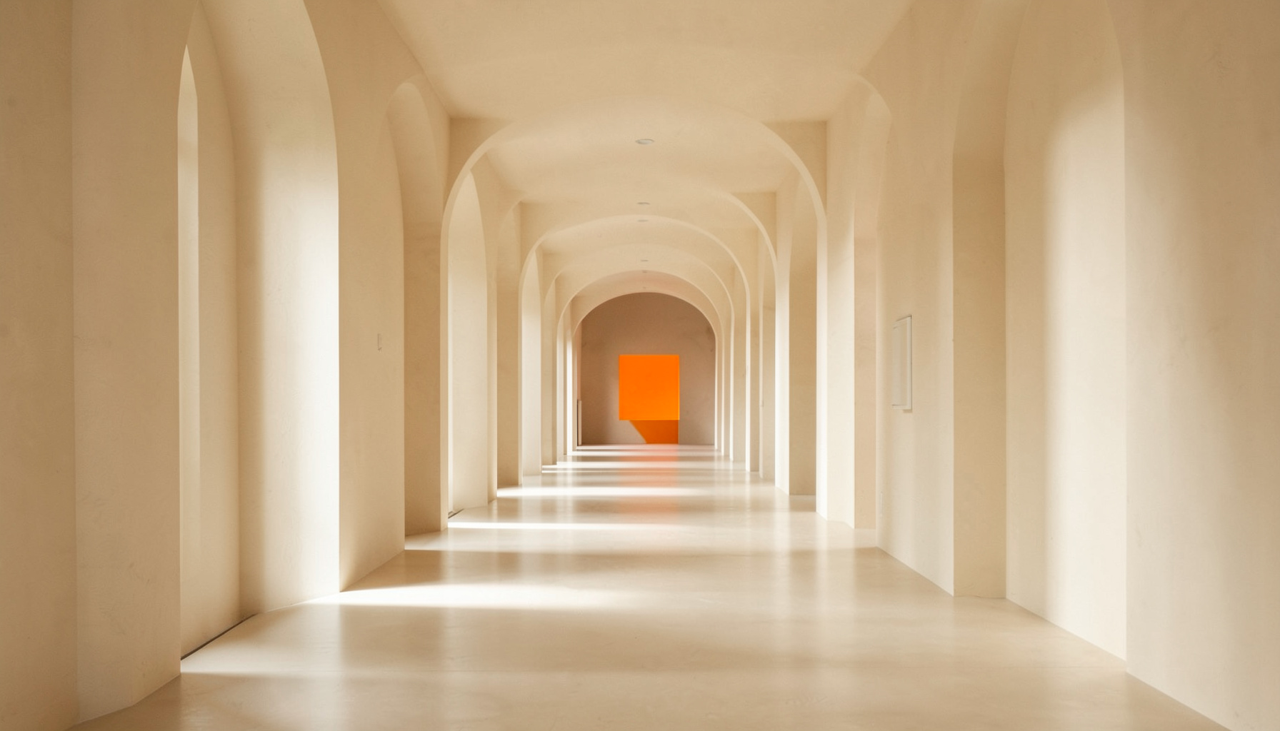 A clean architectural photograph with strong symmetry: a long, quiet corridor with evenly spaced arched columns receding into the distance, soft natural light filtering through, warm beige and cream tones, calm and orderly atmosphere.