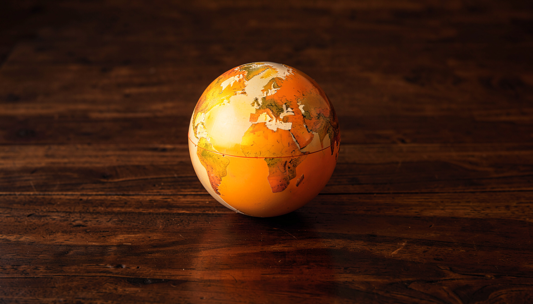 A wide panoramic view of a sunlit globe resting on a dark wooden surface, warm amber and deep brown tones, soft natural light casting gentle shadows across the textured wood grain.