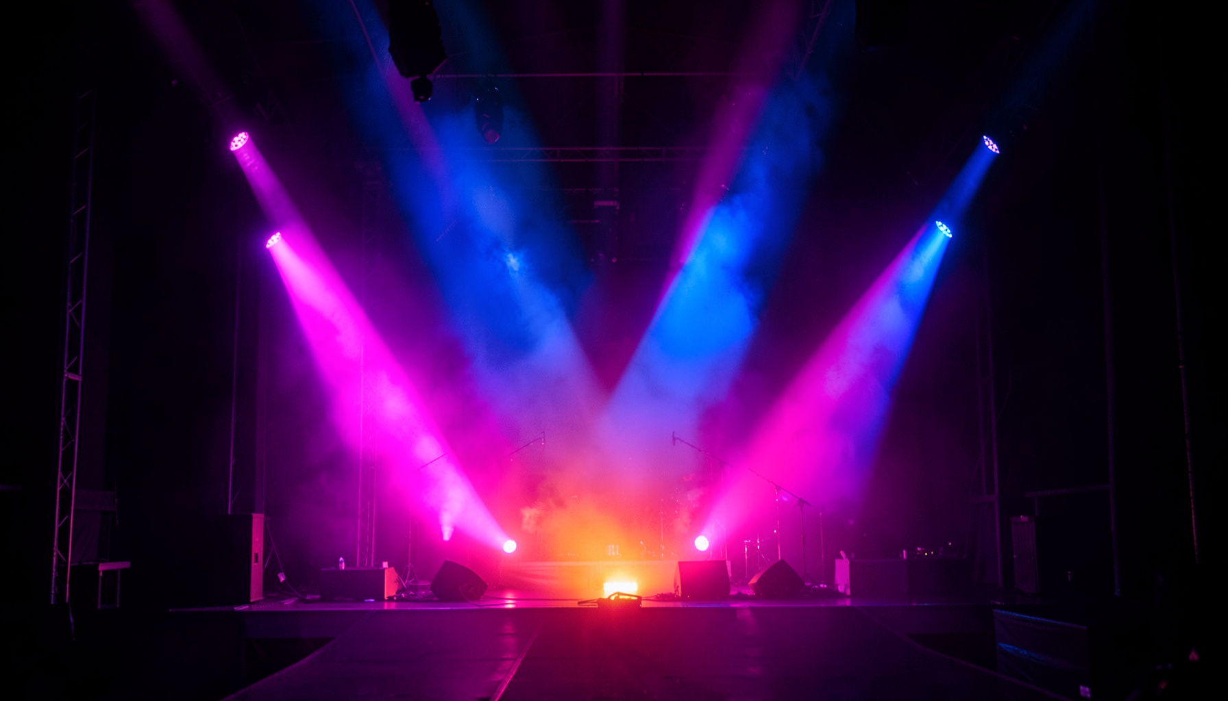 A vibrant concert stage bathed in magenta and electric blue light, with a hazy atmosphere and symmetrical beams cutting through the darkness.