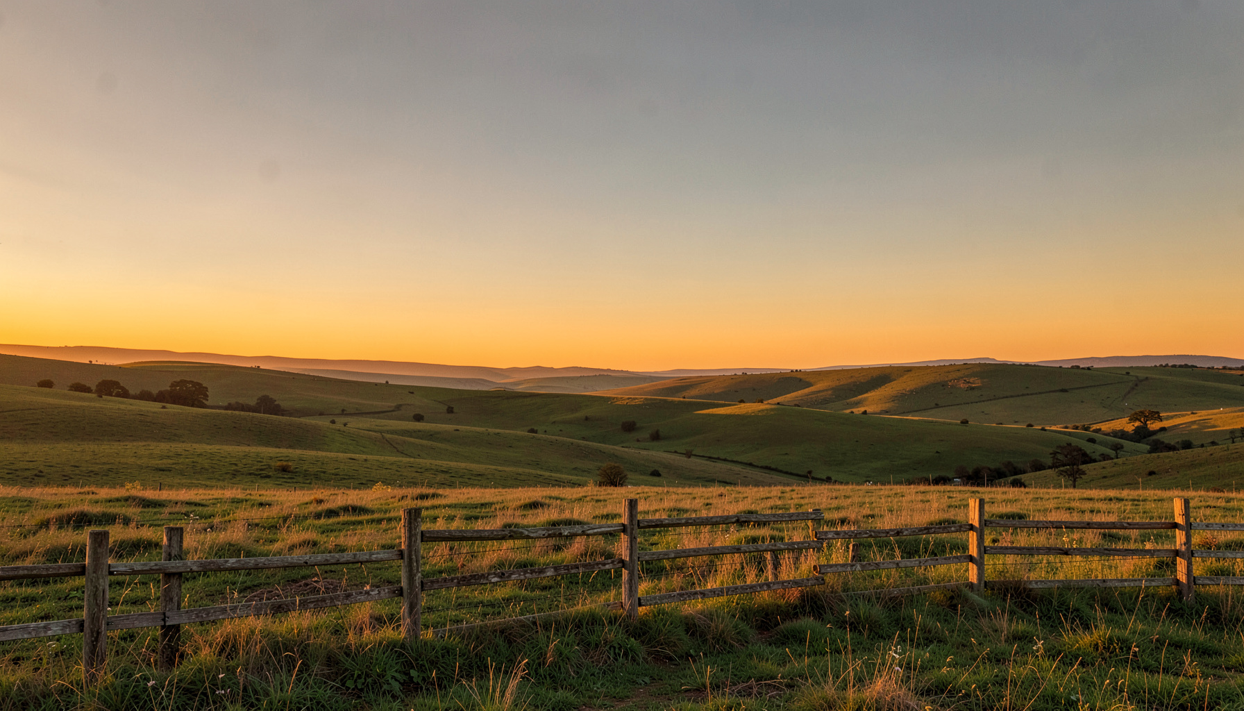 Wide landscape of rolling countryside at golden hour, warm amber and dusty green tones, with a weathered wooden fence stretching across the foreground under a vast open sky.