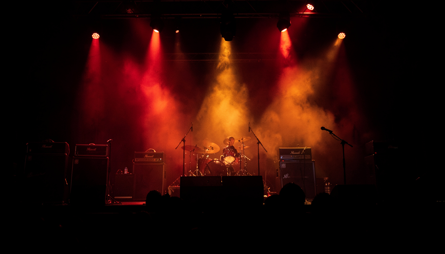 A dark concert stage with dramatic red and amber spotlights cutting through haze, amplifiers silhouetted against the glow, capturing the raw energy of live rock music.